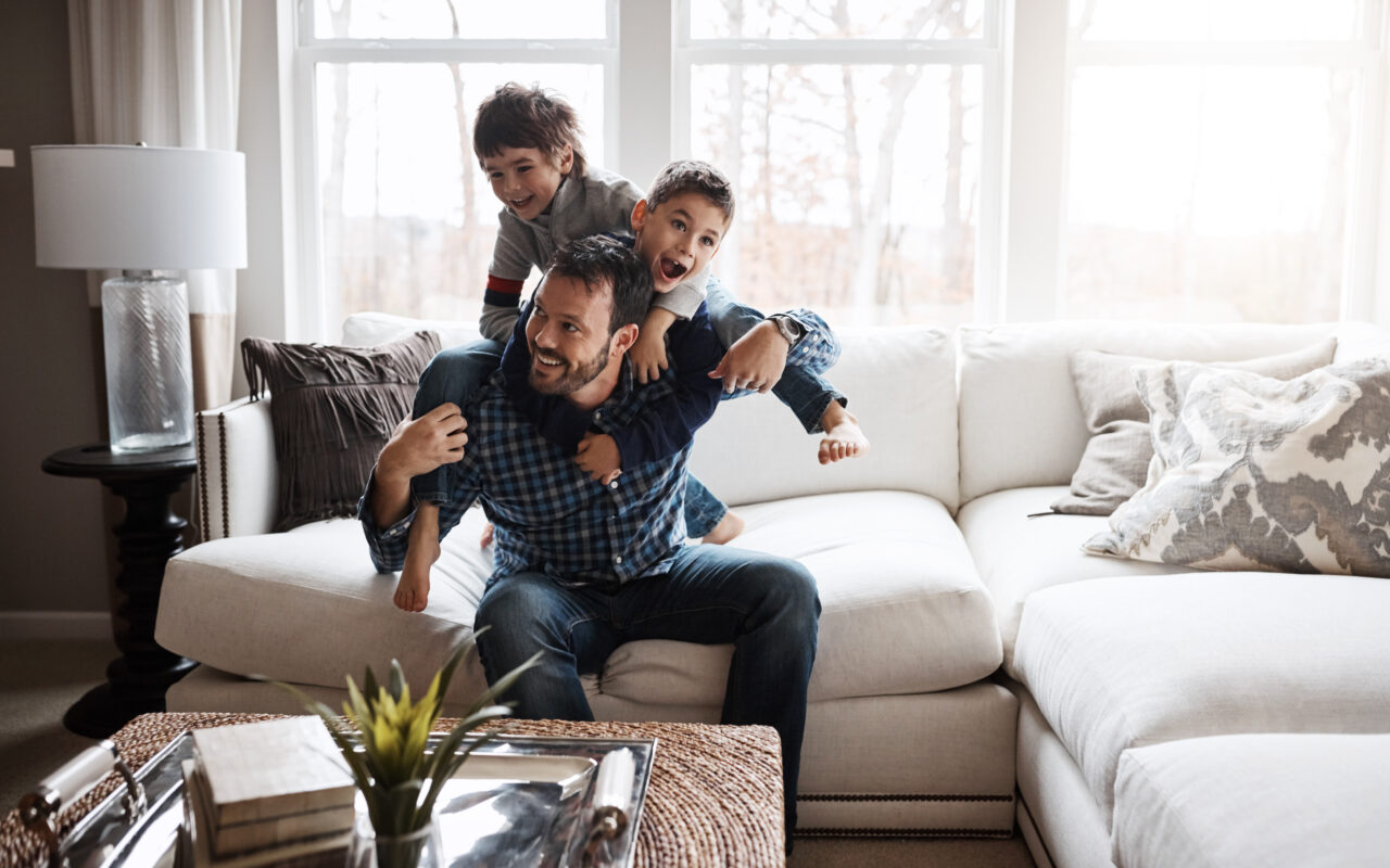 A father playing with two children on a living room sofa.